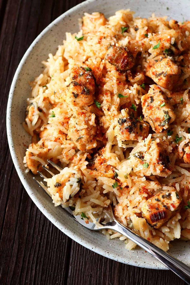 a white bowl filled with rice and meat on top of a wooden table next to a fork