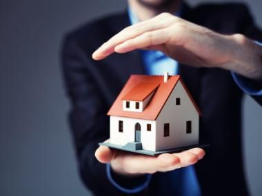 a man in a business suit holding a model house with his hand out to the camera