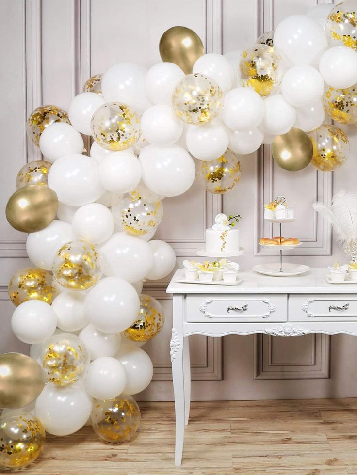 white and gold balloon arch in the corner of a room with a small table, dresser and chair