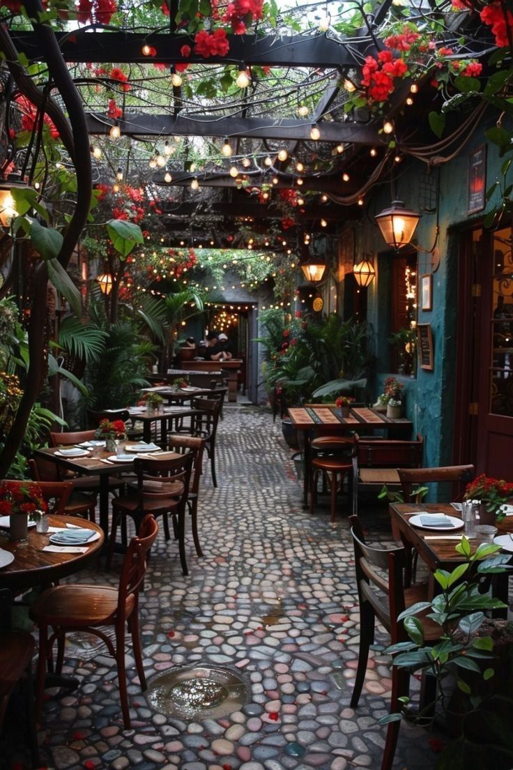 an outdoor restaurant with tables and chairs covered in plants, lights and flowers hanging from the ceiling