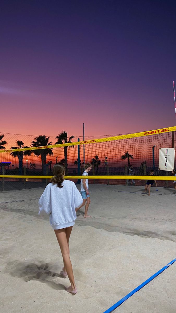 two women are playing volleyball on the beach at sunset or dawn, with palm trees in the background