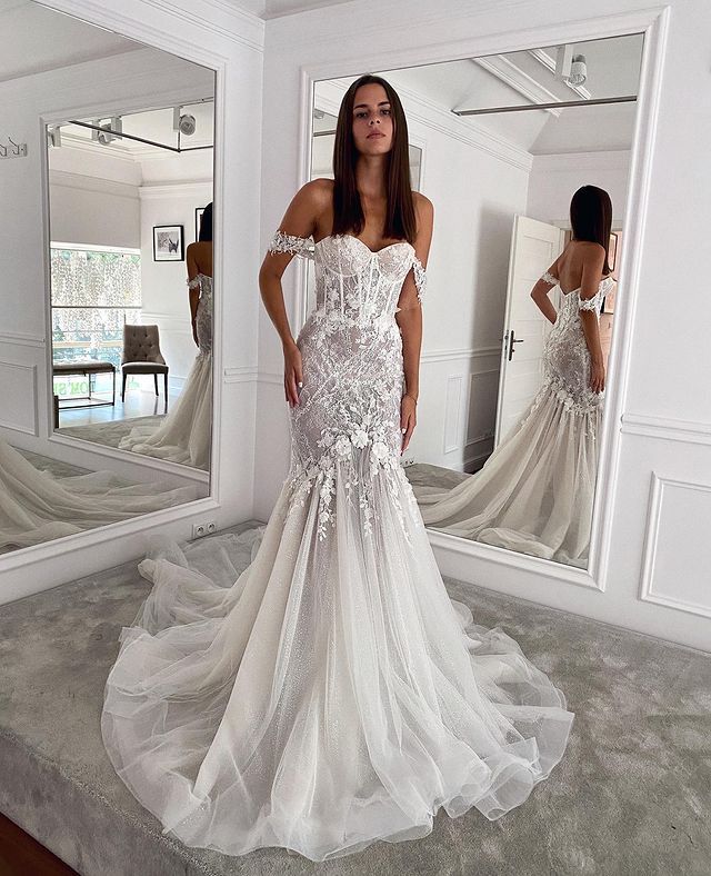 a woman standing in front of a mirror wearing a wedding dress with an off the shoulder design