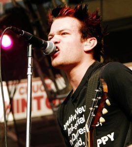 a man singing into a microphone while holding a guitar in his right hand and wearing a black t - shirt with white writing on it