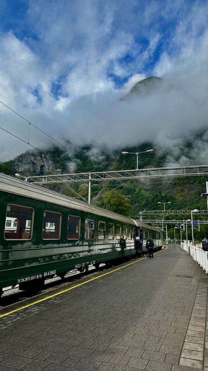 a green train traveling down tracks next to a lush green mountain covered in fog and clouds