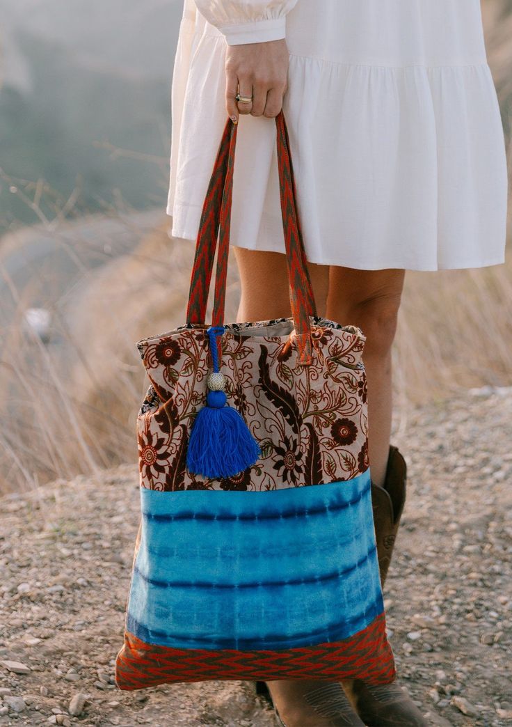 [Color: Natural/Blue] A woman standing outside on a cliff wearing a white dress and holding a bohemian tie dye patchwork tote bag. Featuring a large tassel accent. Bohemian Fabric Shoulder Bag For Daily Use, Bohemian Canvas Bag With Double Handle, Bohemian Multicolor Canvas Bag, Bohemian Canvas Rectangular Bag, Bohemian Fabric Tote Bag, Bohemian Canvas Tote Shoulder Bag, Bohemian Canvas Satchel Bag, Hippie Cotton Shoulder Bag For Travel, Festival Cotton Tote Shoulder Bag