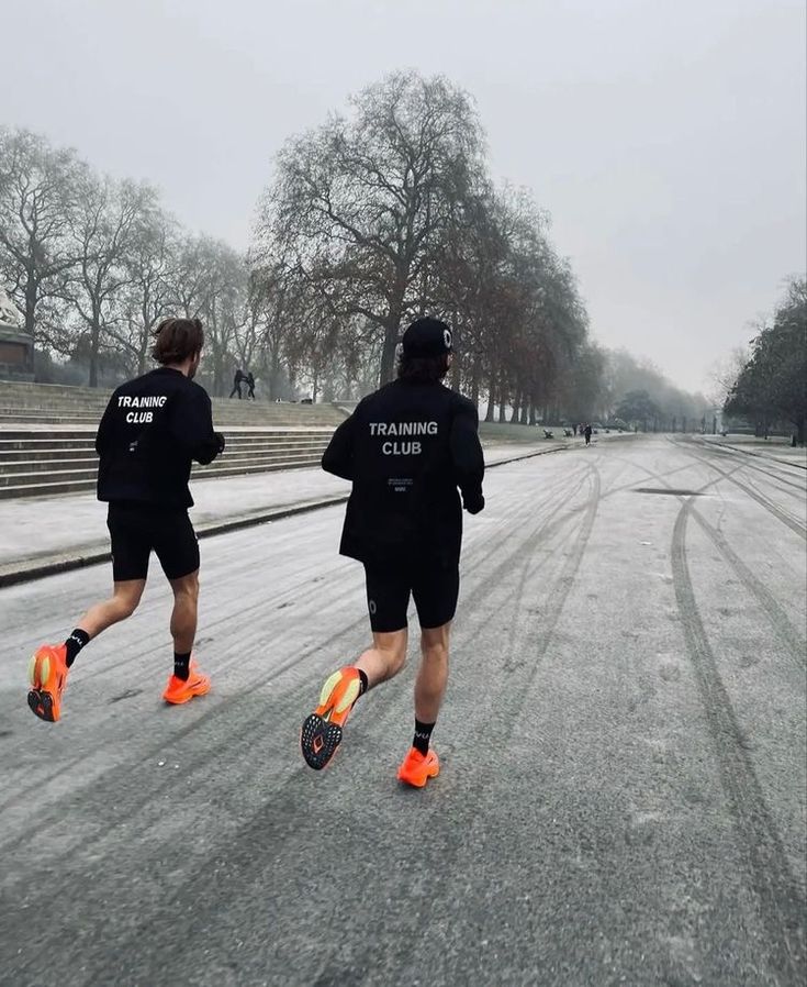 two men in black jackets are running down the street on snow - covered ground with trees and steps behind them