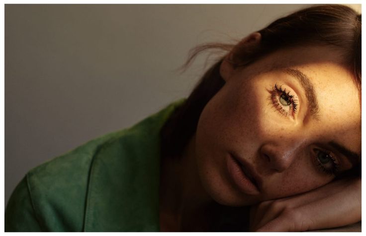 a woman with brown hair and green shirt leaning on a window sill looking at the camera