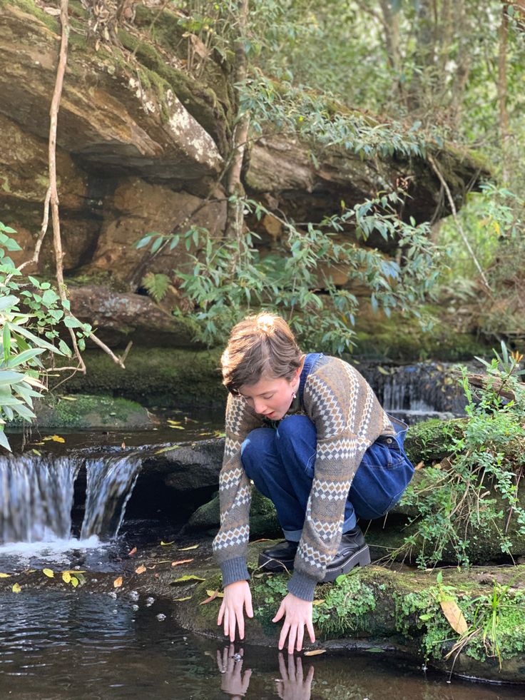 a woman crouches down to look at the water in front of some rocks and trees