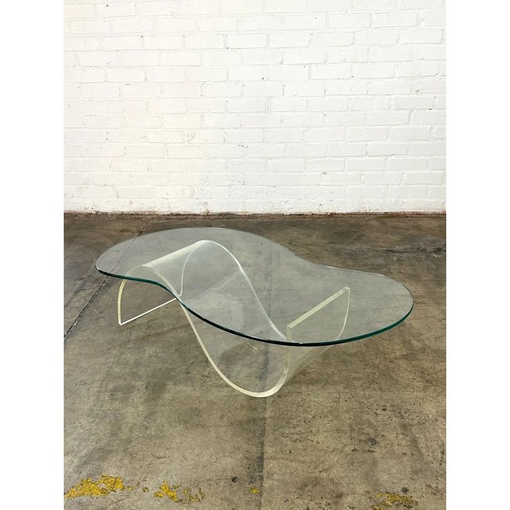 a glass coffee table with curved legs on concrete flooring in front of a white brick wall