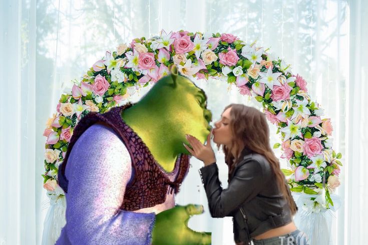 a woman is kissing the face of a green creature in front of a floral wreath