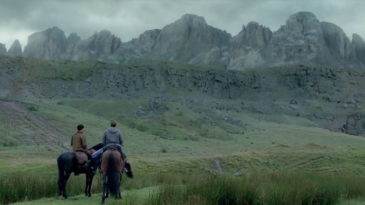 two people are riding horses in front of some mountains and grass, with one person on the horse