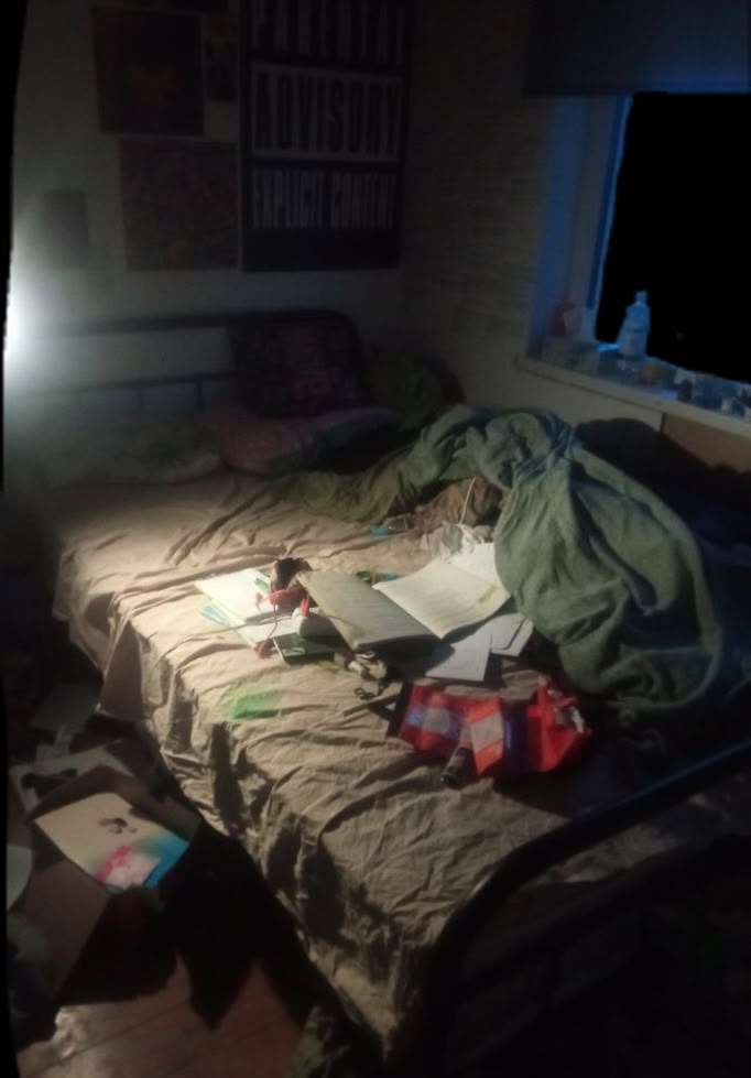 an unmade bed sitting next to a window in a room with lots of clutter