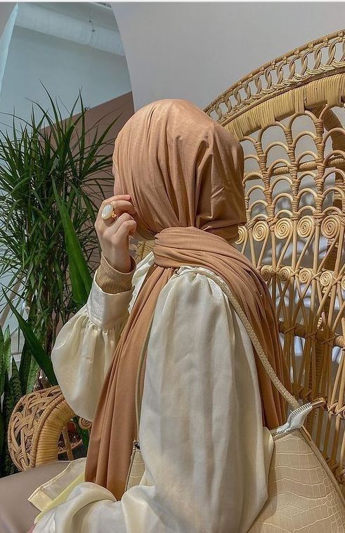 a woman sitting on a wicker chair wearing a beige head scarf and holding a cell phone