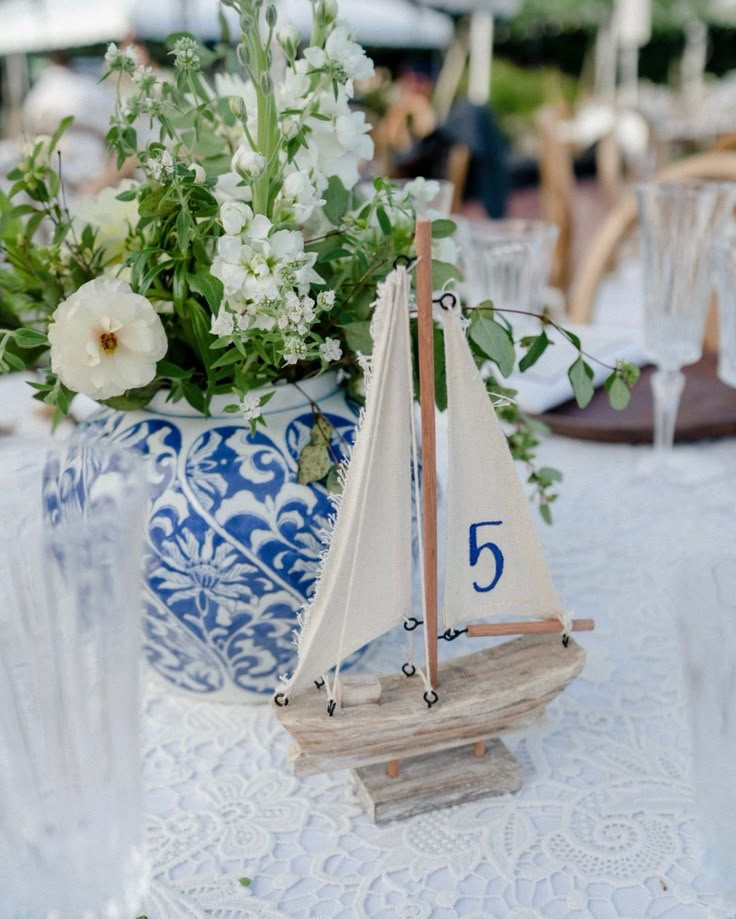 a small sailboat is sitting on a table with flowers in the vase behind it