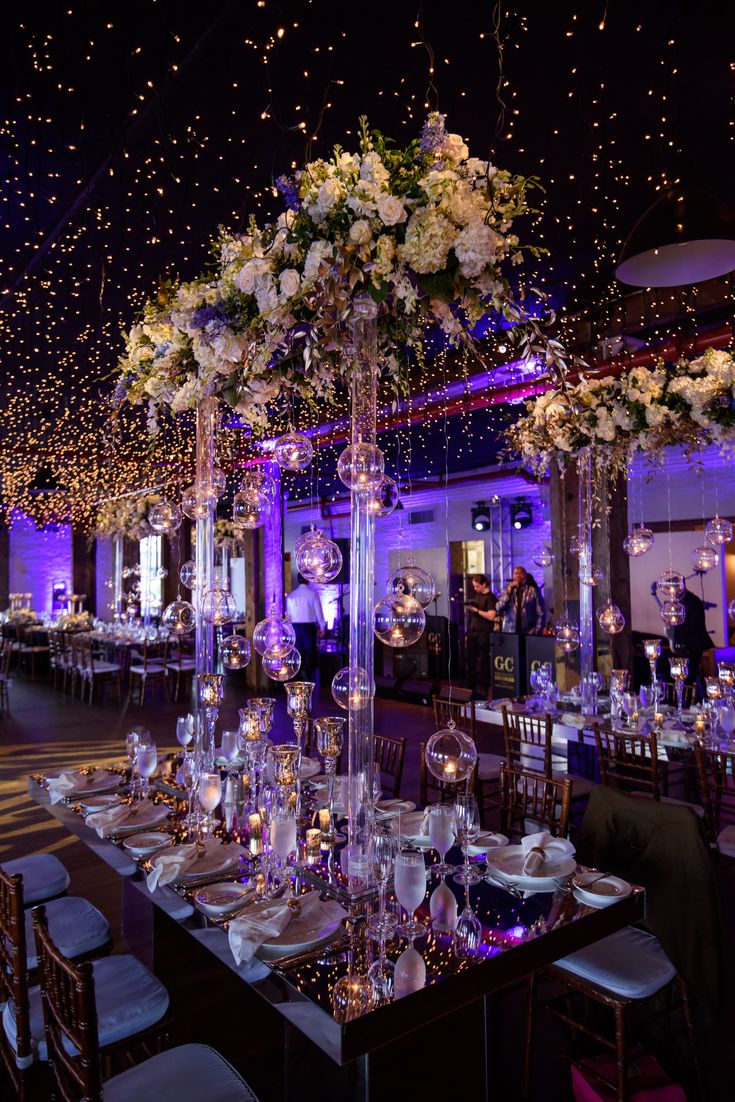 a table set up for an event with white flowers and candles on the centerpieces