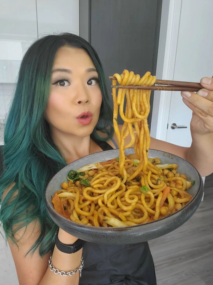 a woman with green hair is holding a plate of noodles and chopsticks in front of her face