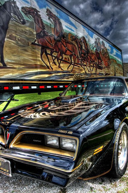 an old car is parked in front of a large trailer with horses on it's side