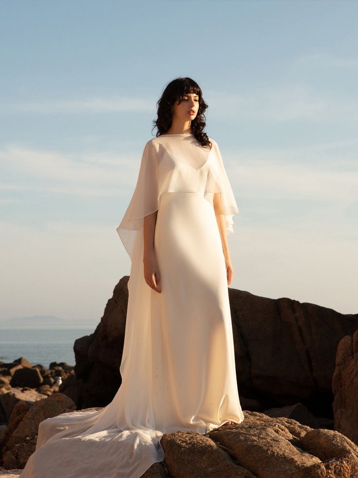 a woman in a white dress standing on rocks near the ocean and looking off into the distance