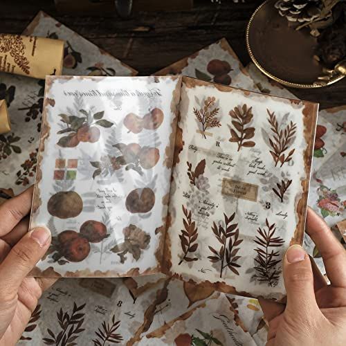 two hands are holding an open book with leaves on it and papers scattered around them