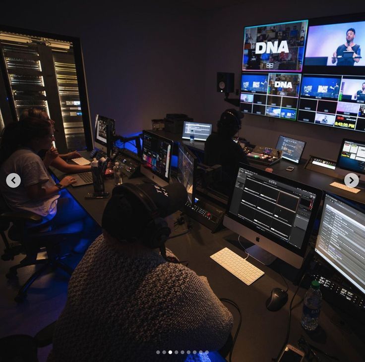 two people sitting in front of multiple computer monitors with video screens on the wall behind them