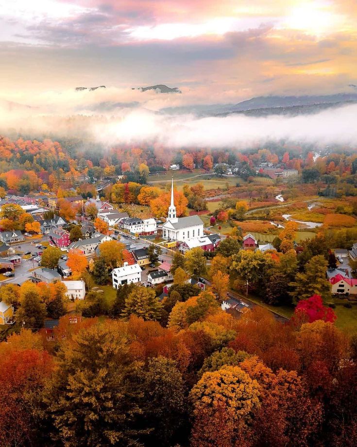 an aerial view of a small town surrounded by autumn trees and fog in the sky