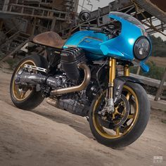 a blue motorcycle parked on top of a dirt road next to a wooden fence and building