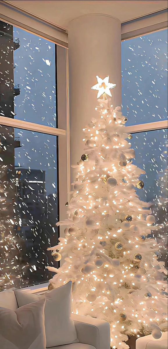 a white christmas tree in front of a window with snow falling down on the ground