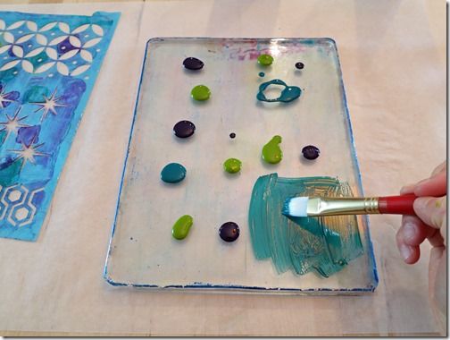 a person is painting on a glass plate with blue and green paint, while another hand holds a brush