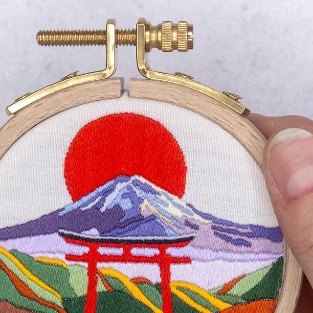 a person holding up a small embroidered object with a mountain in the background and a red sun at the top