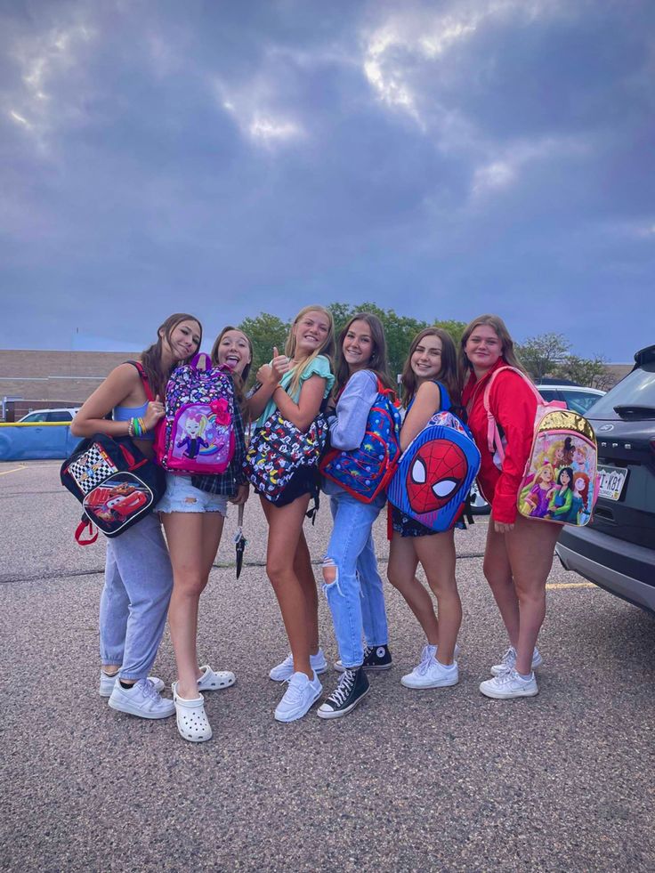 four girls with backpacks are standing in front of a car and posing for the camera