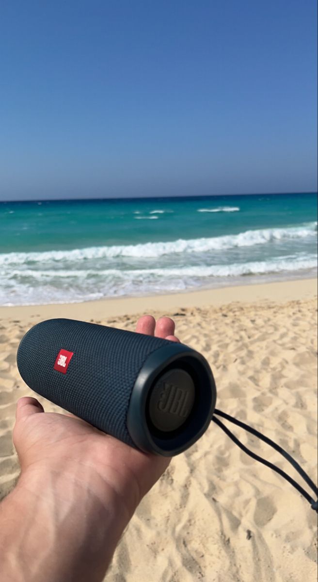 a hand holding a bluetooth speaker on top of a sandy beach next to the ocean