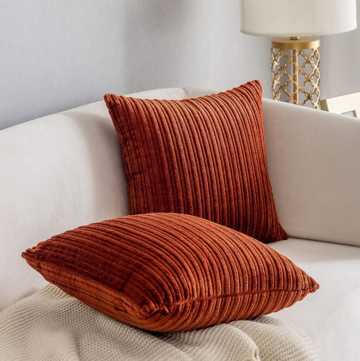 two orange pillows sitting on top of a white couch next to a lamp and pillow case