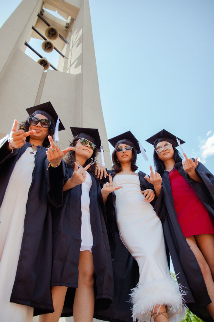 four young women in graduation gowns posing for the camera with their thumbs up and one wearing sunglasses