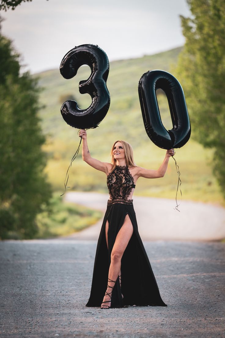 a woman in a long black dress holding up the number thirty three balloons while standing on a road