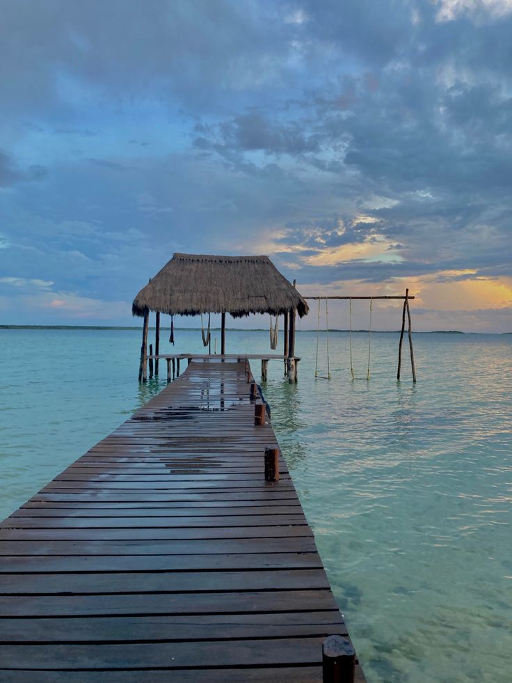 a wooden dock leading into the ocean with a thatched roof over it at sunset