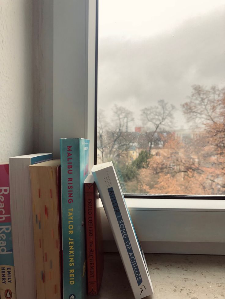 two books sitting on top of each other next to a window sill with trees outside