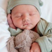 a baby sleeping with a stuffed animal in it's lap and wearing a green hat