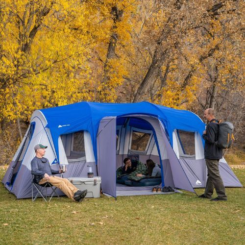 two men standing next to a tent in the grass with trees behind them and one man sitting on a chair