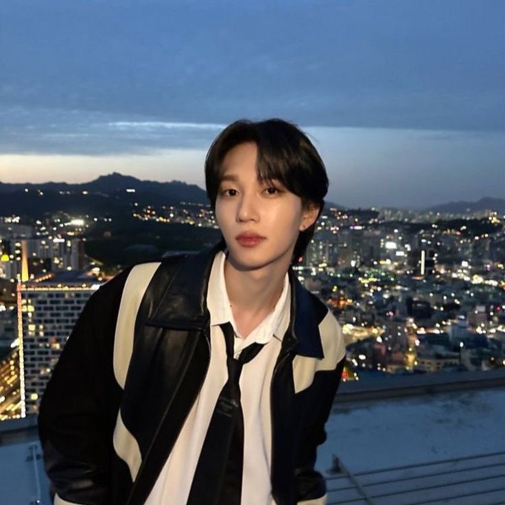 a man standing on top of a building next to a city at night with his hands in his pockets