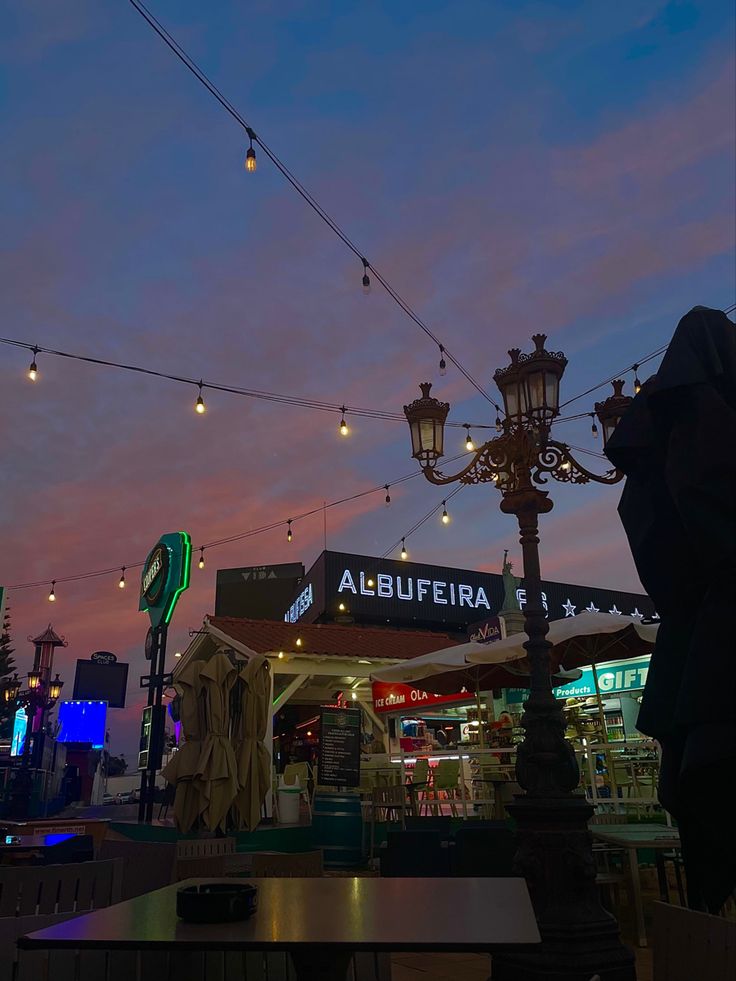 an outdoor restaurant with lights strung from the ceiling and tables in front of it at dusk