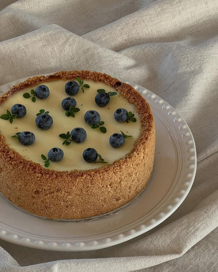 a cheesecake with blueberries on top is sitting on a white plate and sits on a beige cloth