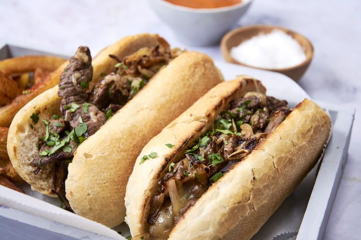 two sandwiches with meat and onions in a tray next to french fries on a table