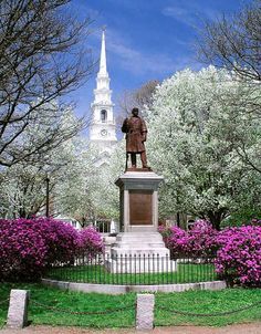 a statue in the middle of a park with trees and flowers around it, next to a church steeple