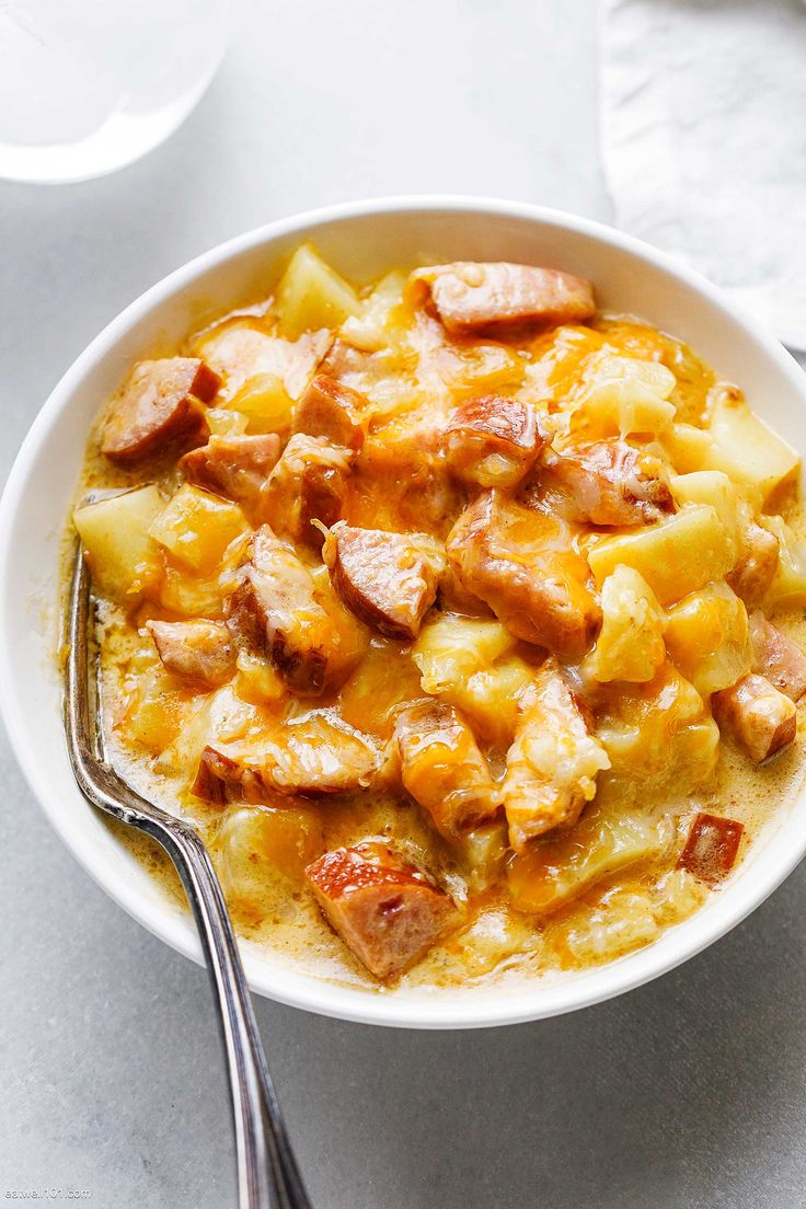 a bowl filled with potatoes and sausages next to a fork on a white surface