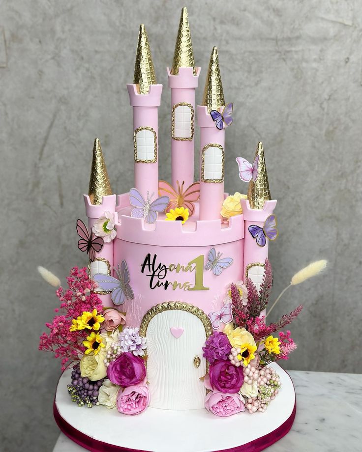 a pink castle cake with flowers and butterflies on it's side, sitting on top of a table