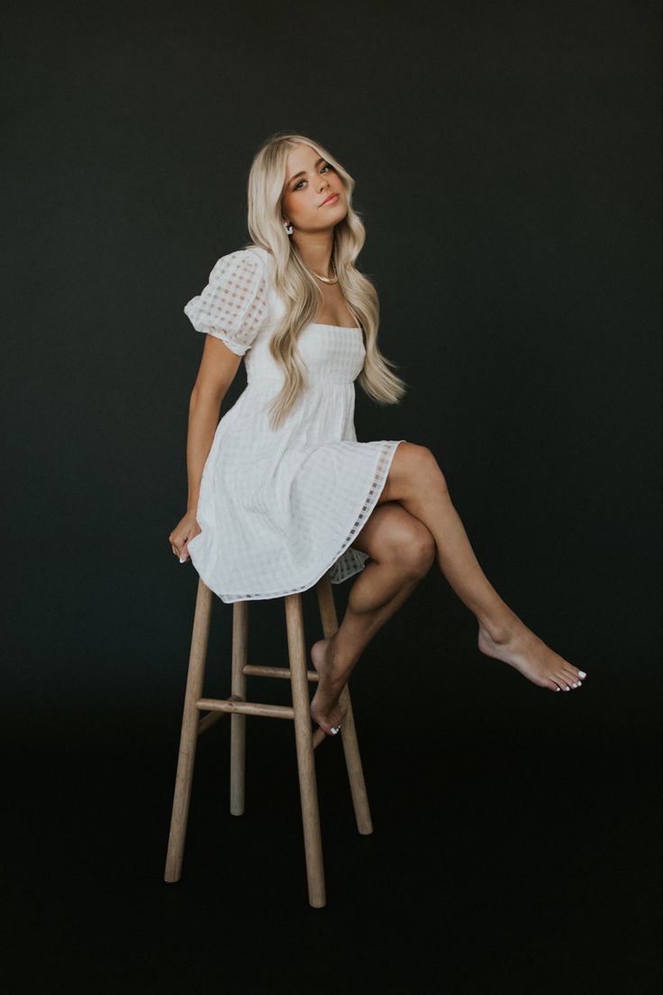 a woman sitting on top of a wooden stool wearing a white dress and high heels