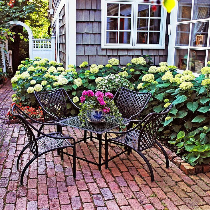 an outdoor table and chairs with flowers in the center on a brick path next to a house