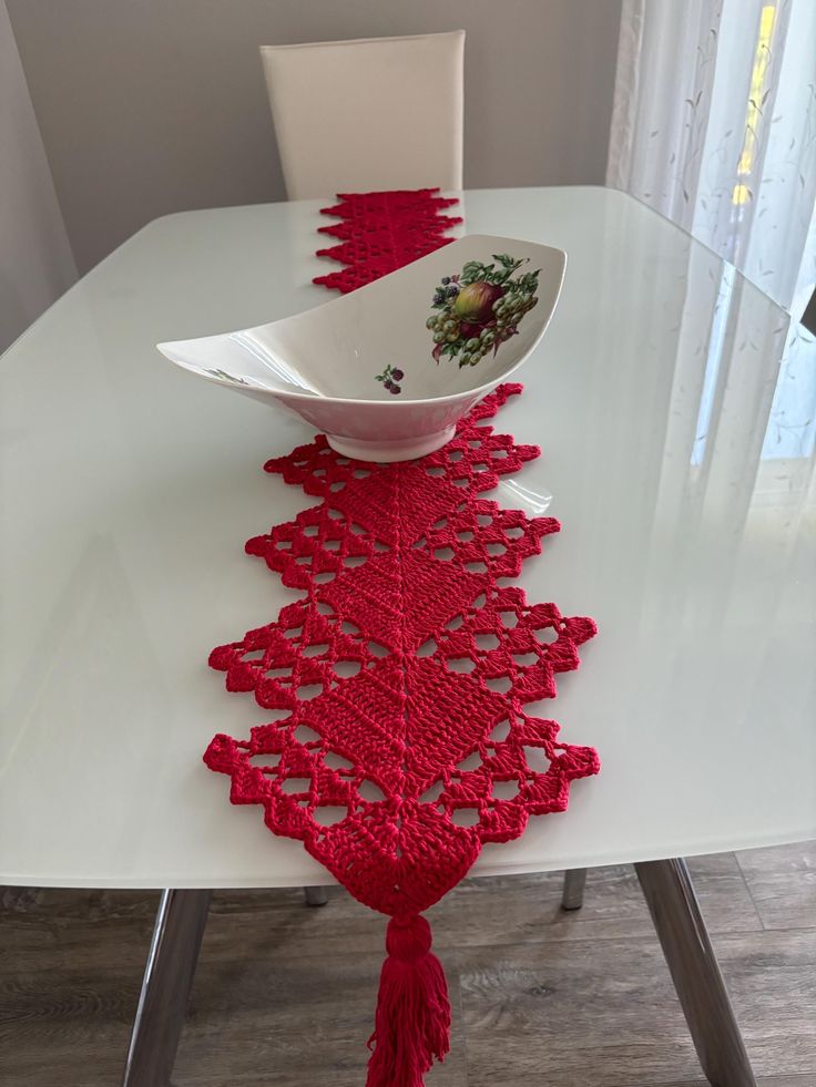 a white table topped with a bowl and red tasselled placemat on top of it