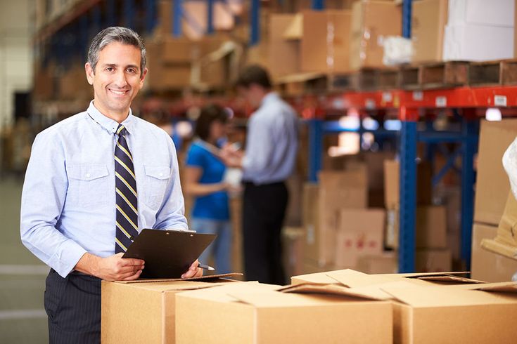 a man standing in front of boxes looking at something on his clipboard while another man stands behind him