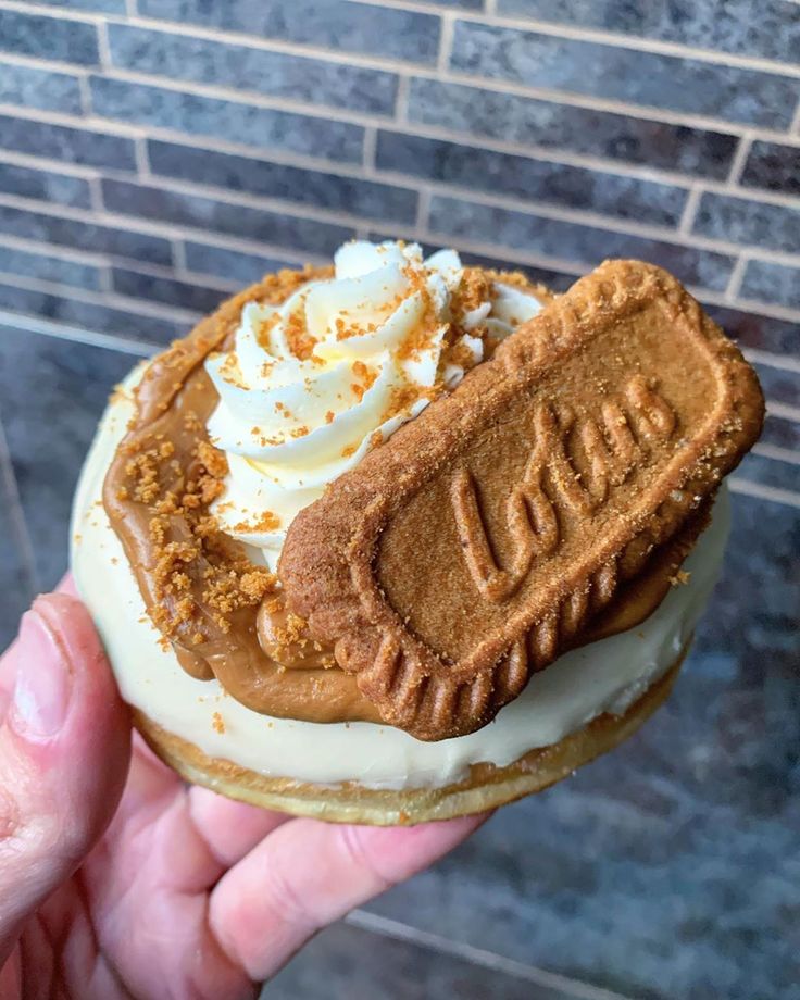 a hand holding a pastry with whipped cream and cookies on top, in front of a brick wall